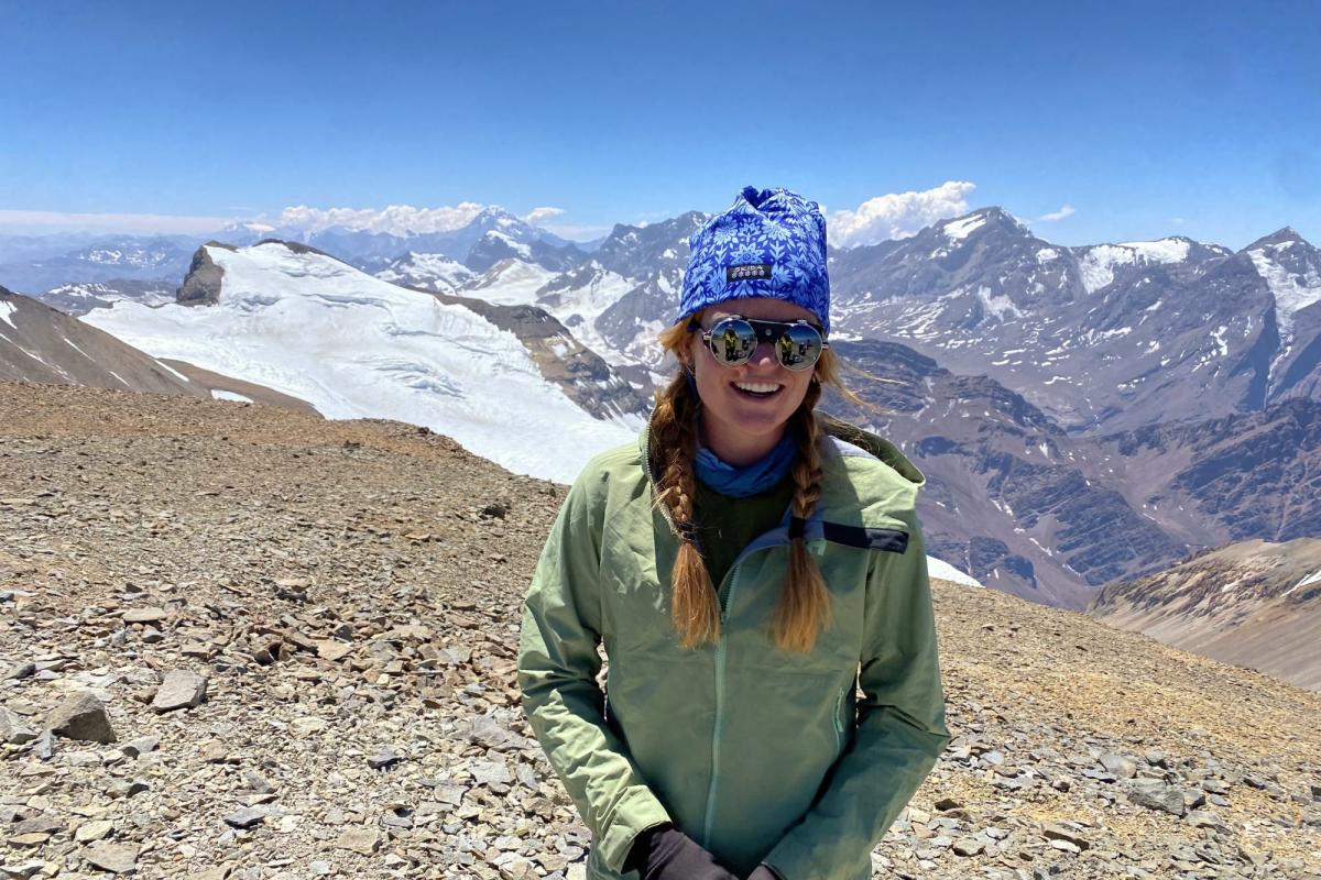 Millie Spencer smiles while standing on the sunny summit of Cerro El Plomo (17,795 feet) in Central Chile. Behind the rocky summit is a prominent glacier, with a background of mountain peaks with more glaciers and snowfields. She is wearing a green jacket, blue knit hat, and mirrored glacier glasses