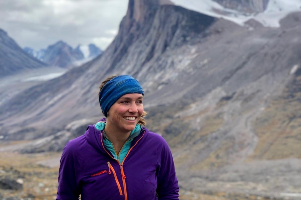 Sarah Crump, in purple coat, stands in front of a dramatic Baffin Island landscape of colorful tundra suddenly broken by towering cliffs.