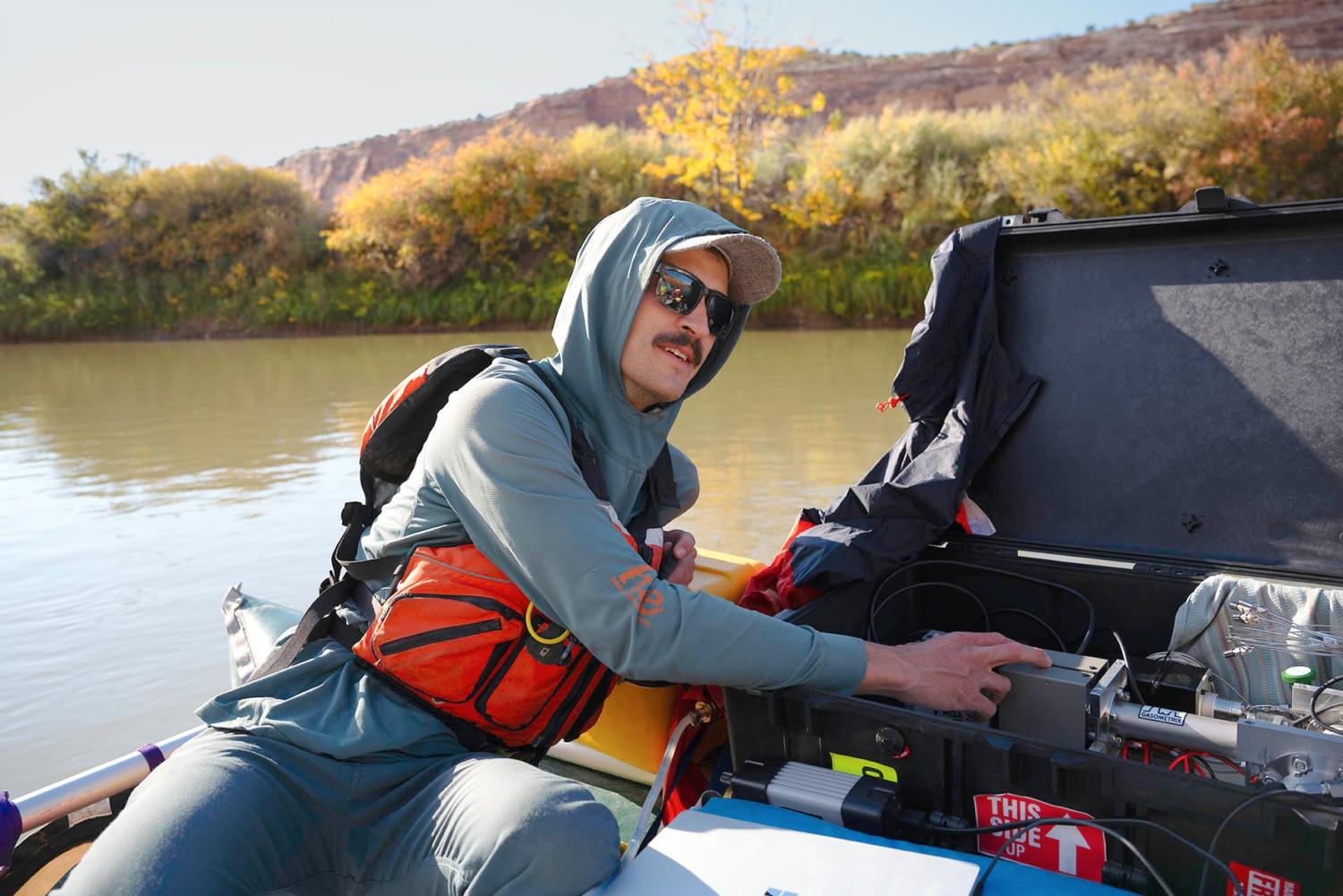 Connor Newman - in sunglasses, hoodie, and orange safety vest - leans over a scientific gadget while on a raft-based survey of water quality on the upper Colorado River.