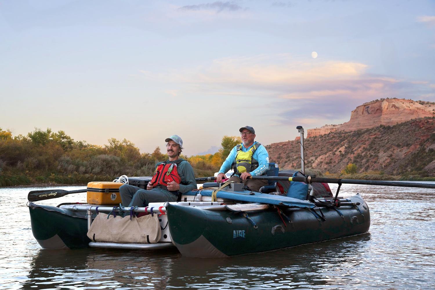 As darkness sets, two people sit in a fully loaded inflatable raft on the upper Colorado River. A nearly full moon rises in the sky above a light colored cliff.