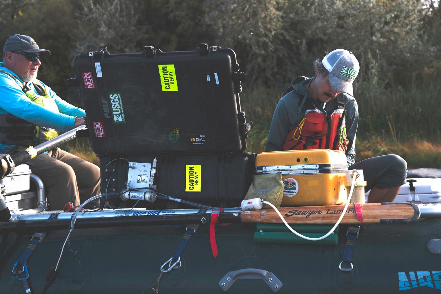 Mike Gooseff and Conor Newman work from a fully packed raft while surveying water quality on the upper Colorado River. Between them are large plastic boxes with tubes and scientific sampling and measuring equipment.