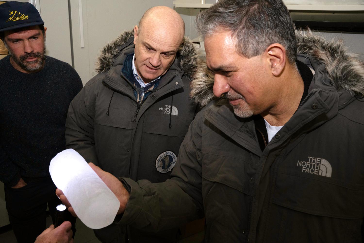 Three people in dark-colored puffy coats look at a section of an ice core with a flashlight shining through it
