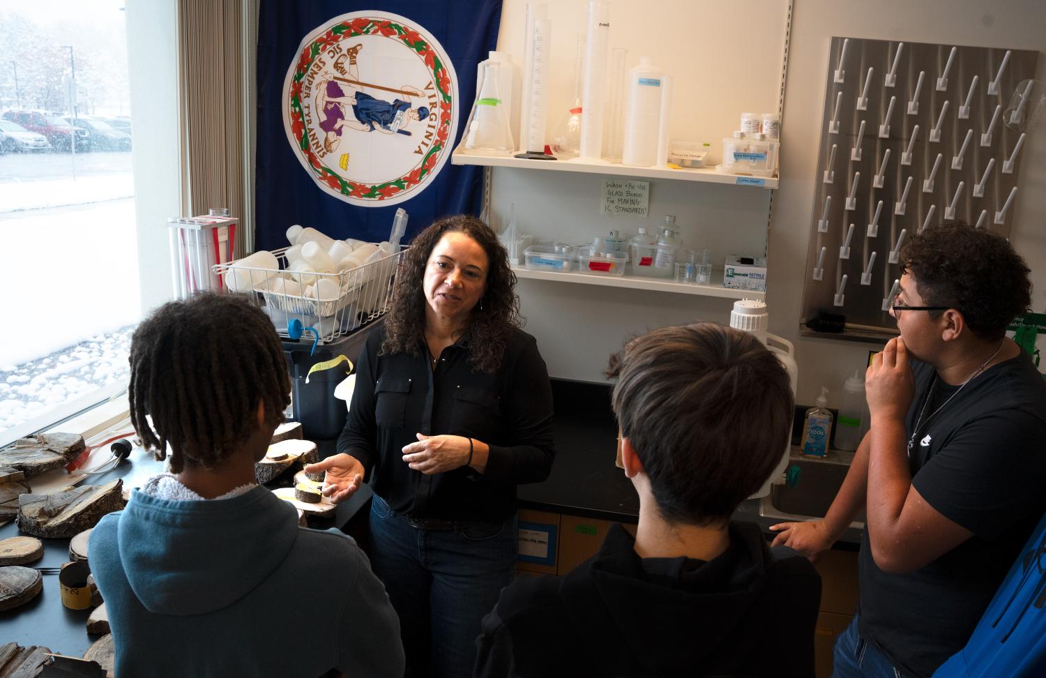 Three young teens listen to a middle-aged woman in a black sweater as she holds a slice of a small tree