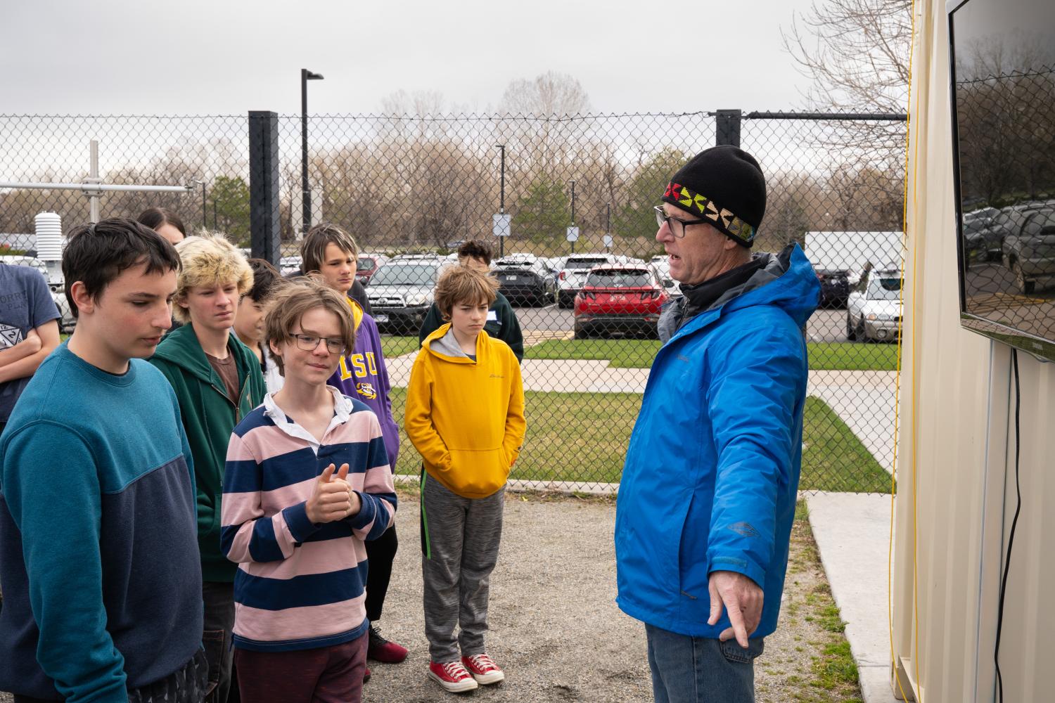 A man in a blue coat points off screen while talking to a group of tweens in a fenced in area outside on a cloudy day