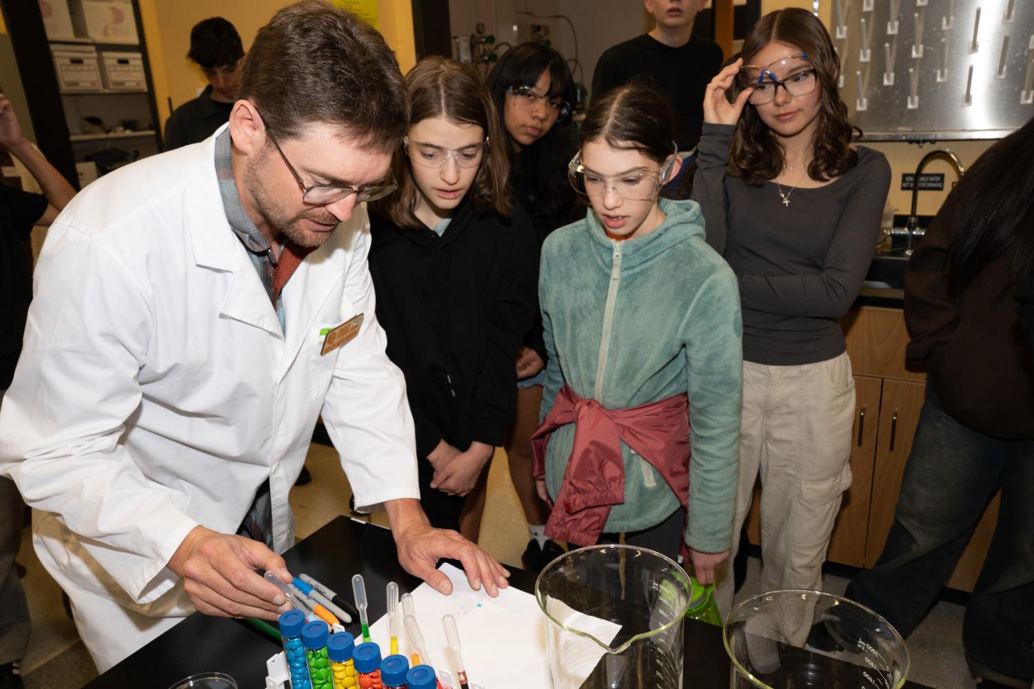 A man in a lab coat drops food coloring onto paper as tweens look on with interest