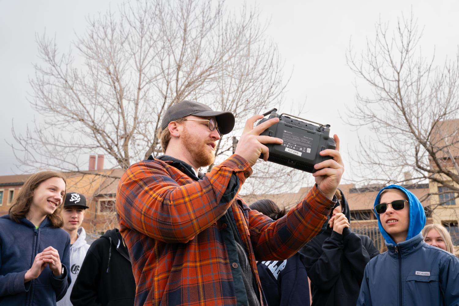 A man in a flannel holds a drone controller while tweens look on with interest outside on a cloudy day