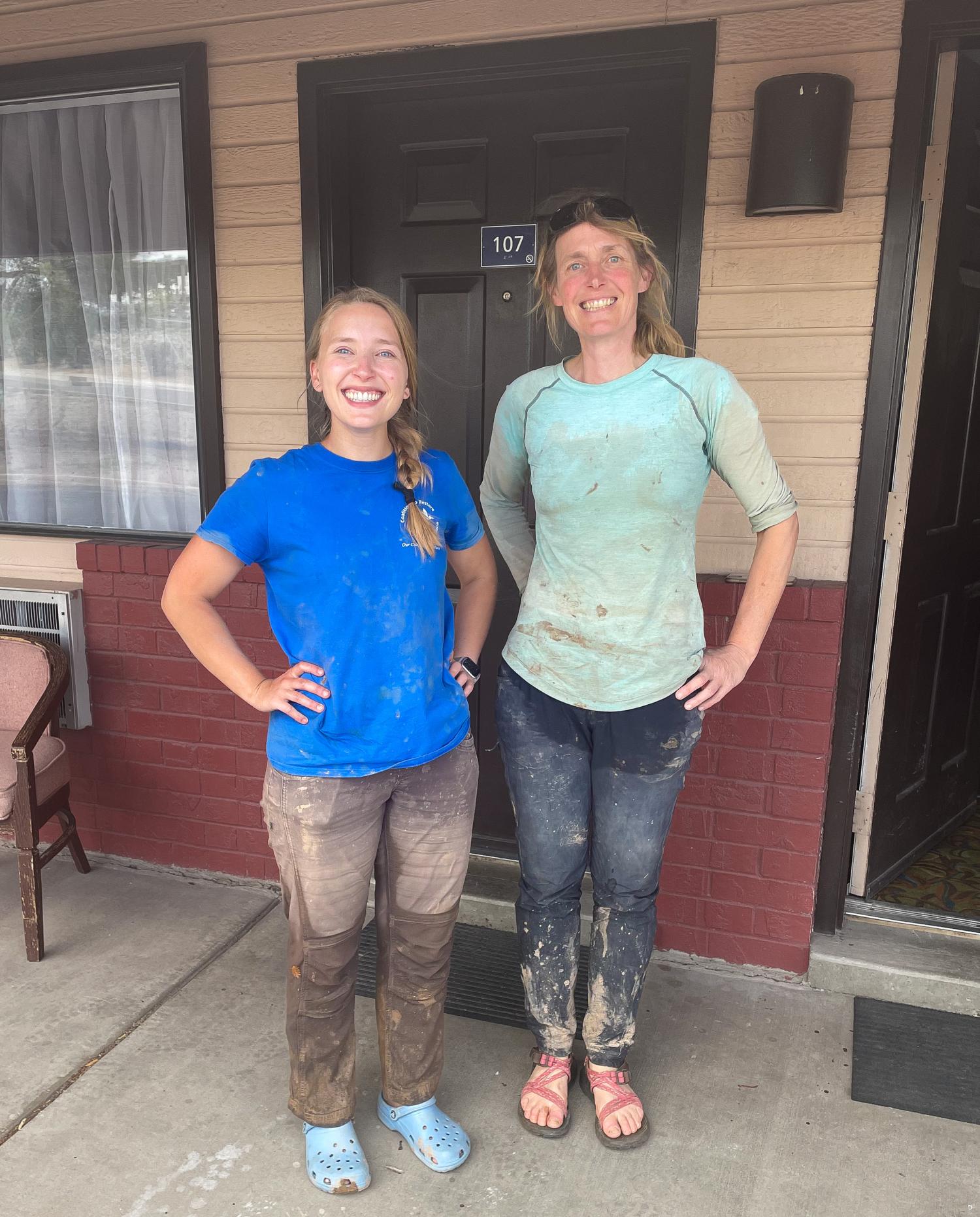 two blonde female scientists, one early-middle-aged and one a young adult, smile in muddy closthes in front of a gray door