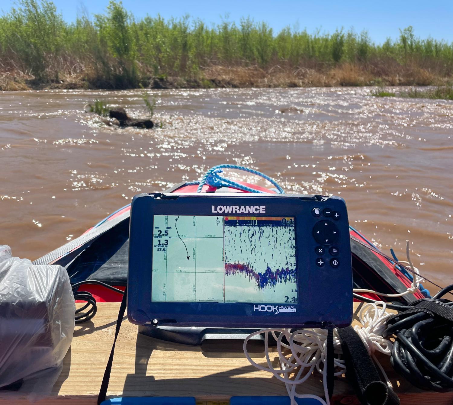 A monitor strapped to the front of a raft in a muddy river displays a spatial chart with dense data points