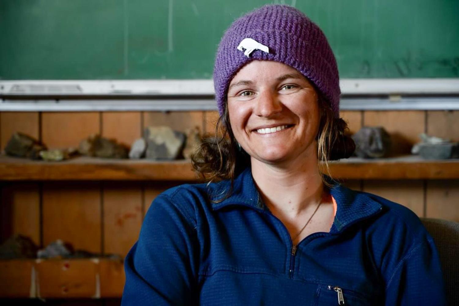A young brunette woman in athletic wear smiles for the camera while seated in front of a rock collection and a green chalkboard