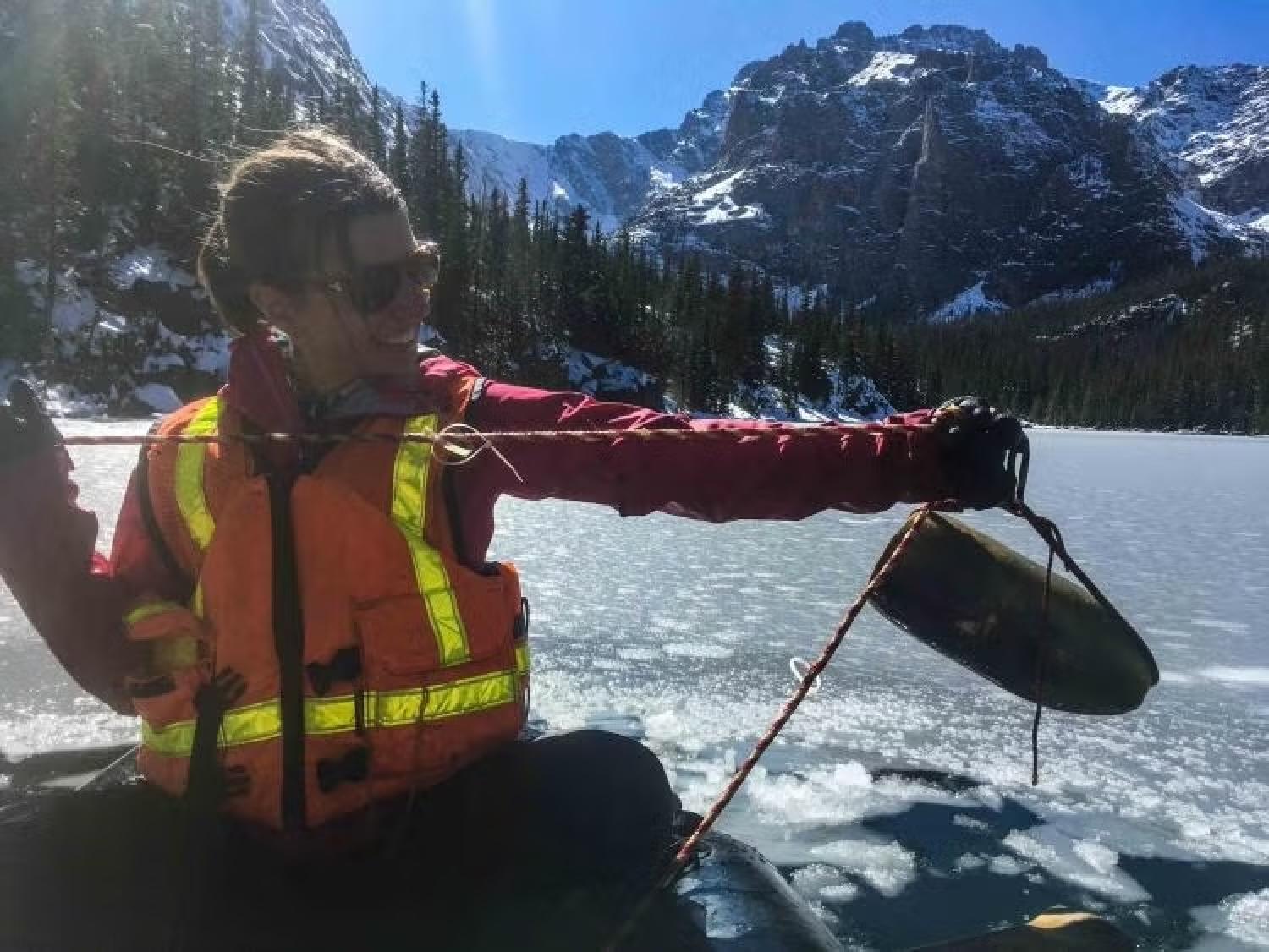 A woman in an orange vest measures something atop a one-person raft in an alpine lake