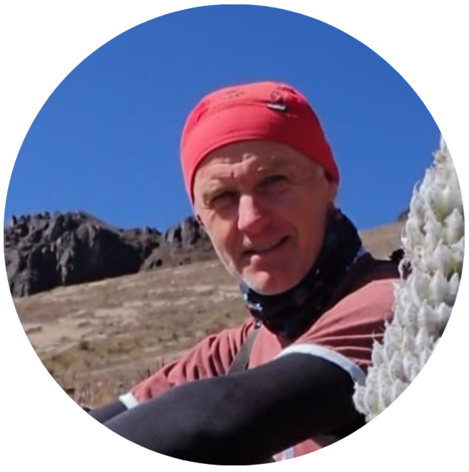Petr sitting on a hillside with rock formations behind him and mountain flora on the right side of the photo.