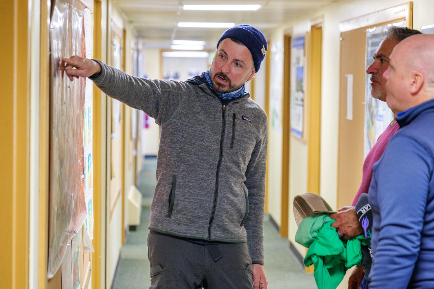 A man points to a map on a wall in a hallways with warm lighting while two other man look on