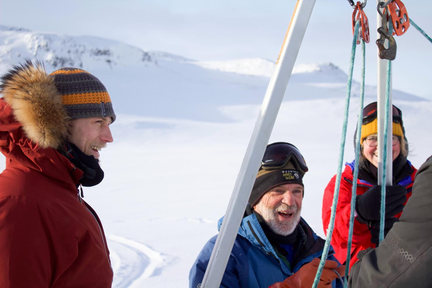 Three researchers in brightly-colored winter gear mess with a complex of metal beams and cord against a backdrop of snow-covered rocky terrain
