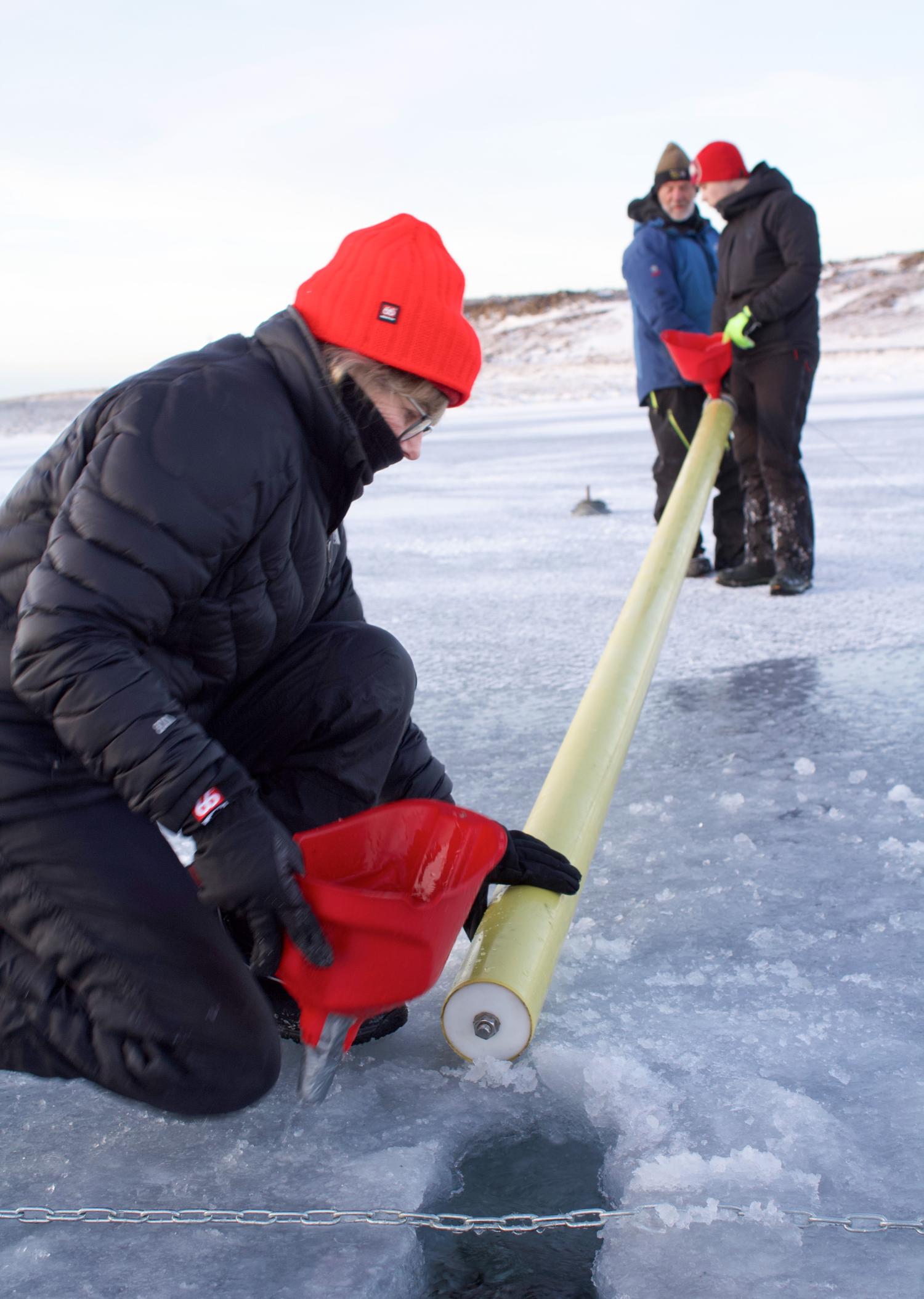 A researcher in a bright red beanie kneels beside a hole in the ice with a hand on a long, yellow metal tube that two other researchers hold on the other end in the background
