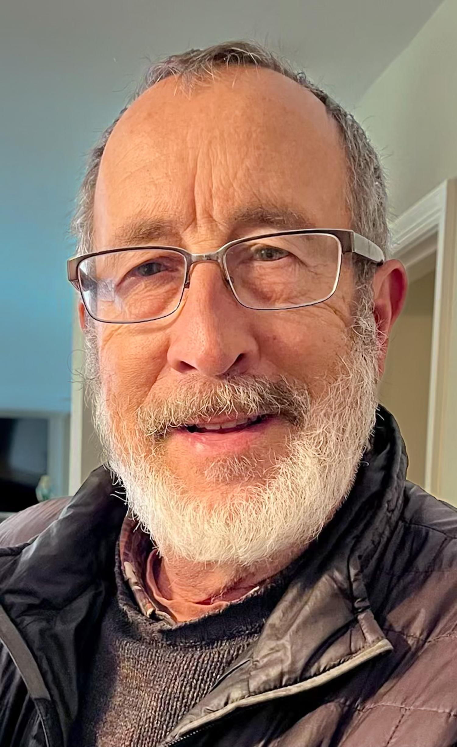 A man with a trimmed white beard in glasses, a dark sweater and an unzipped puffy coat poses for a portrait. Cropped close