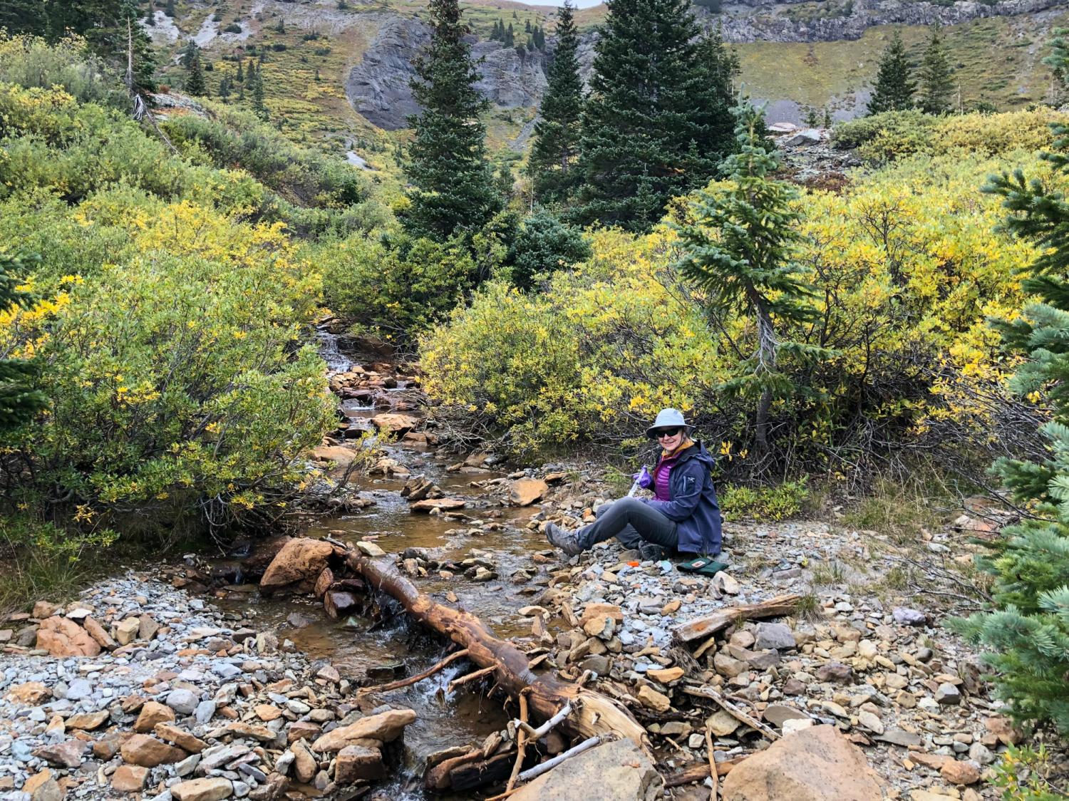 A woman in outdoor clothing sits by a mountain creek with sampling instruments in her hands