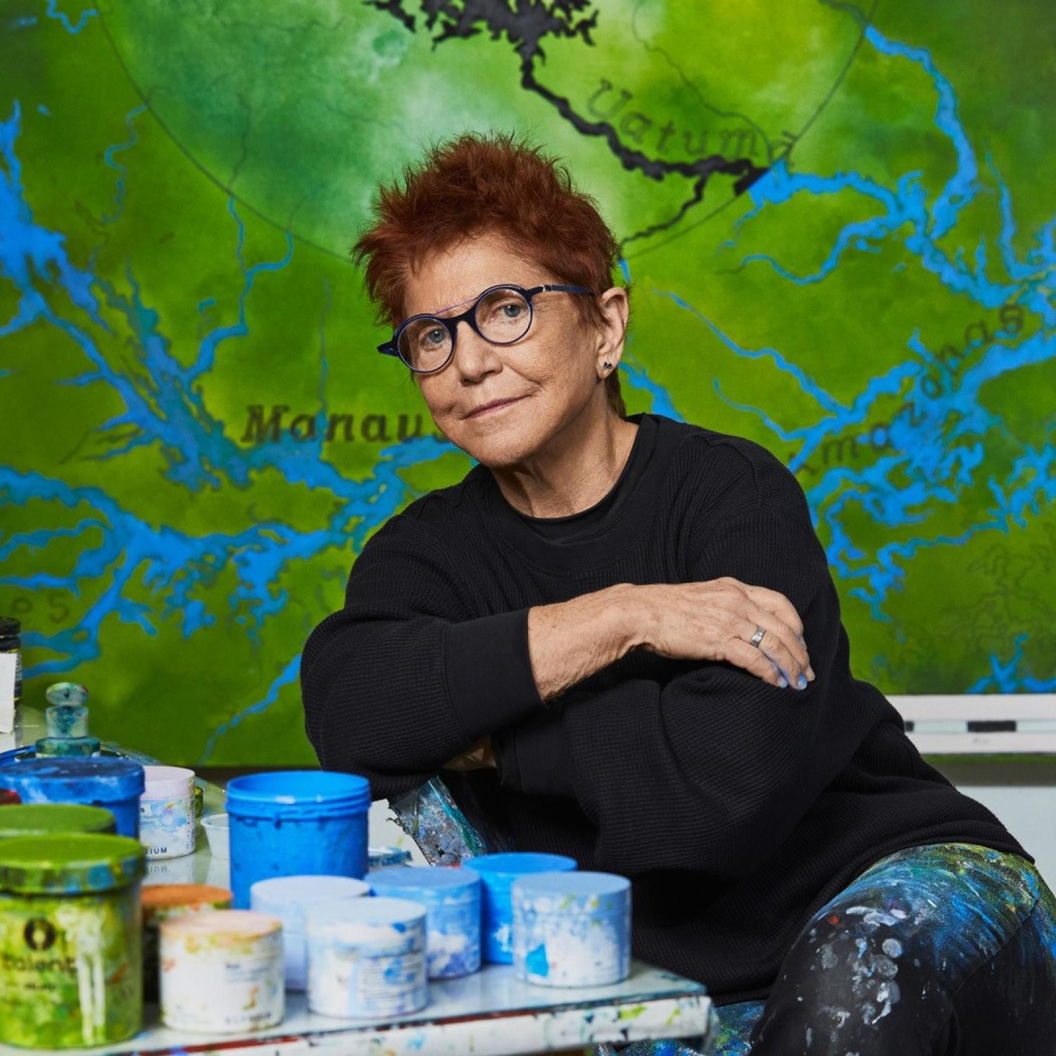 Diane Burko in her studio with a map behind her. She is leaning on a table covered in paint and paint cans