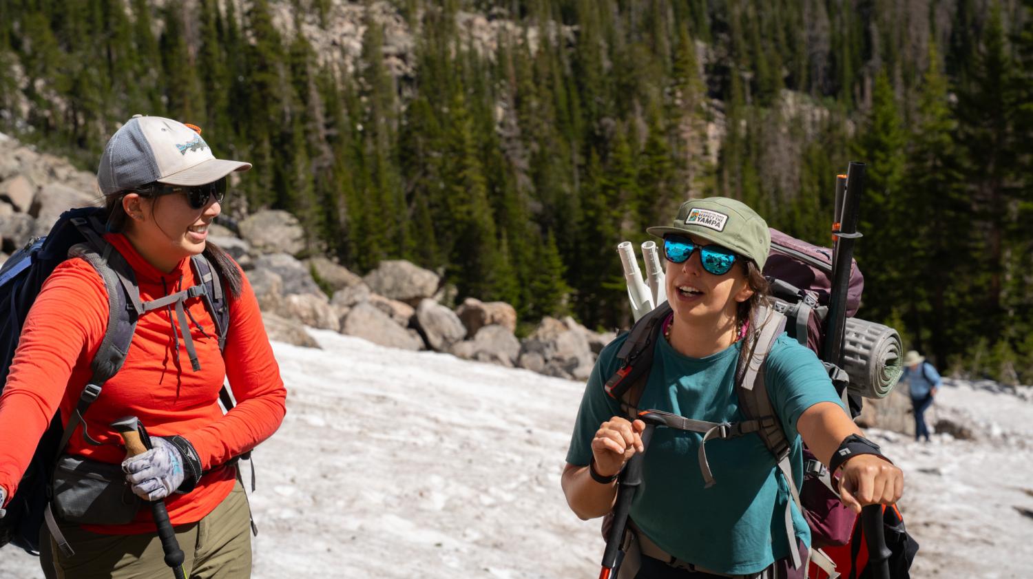 Two women in hiking gear and large backpacks chat atop a steep snowfield with boulders and pine trees in the background
