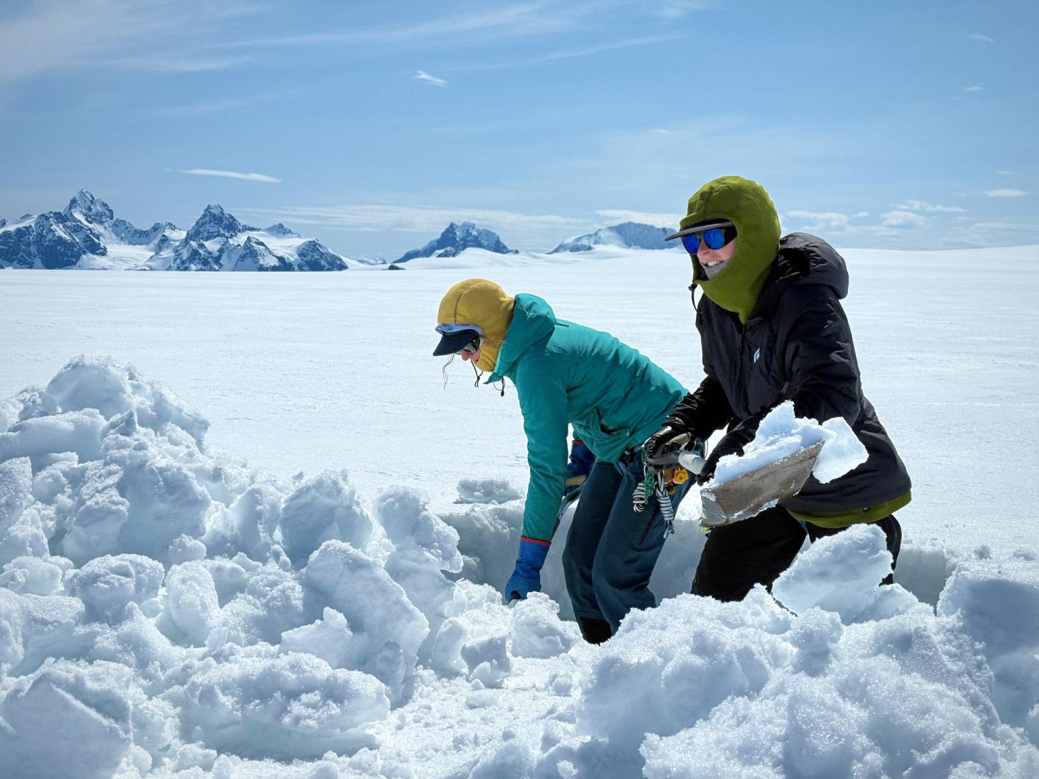Two women in colorful warm outdoor clothing shovel snow out of a shallow pit on a vast expanse of flat snow with mountains in the background