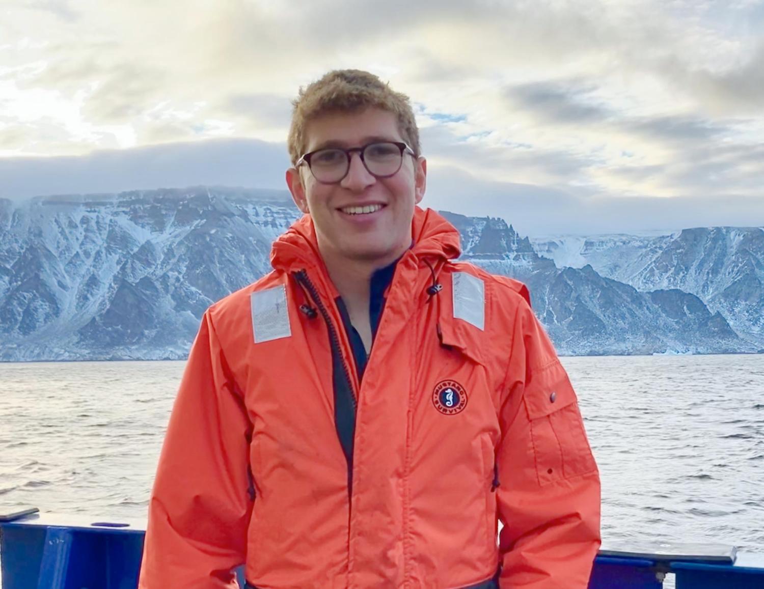 Jed Lenetsky, in orange jacket, stands at a ship's rail with snowy rocky cliffs behind him