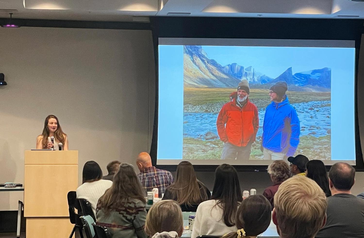 Katie Gannon speaking at the 2024 INSTAAR celebration luncheon, with people seated at tables and a projector screen showing Giff Miller and Sarah Crump on Baffin Island