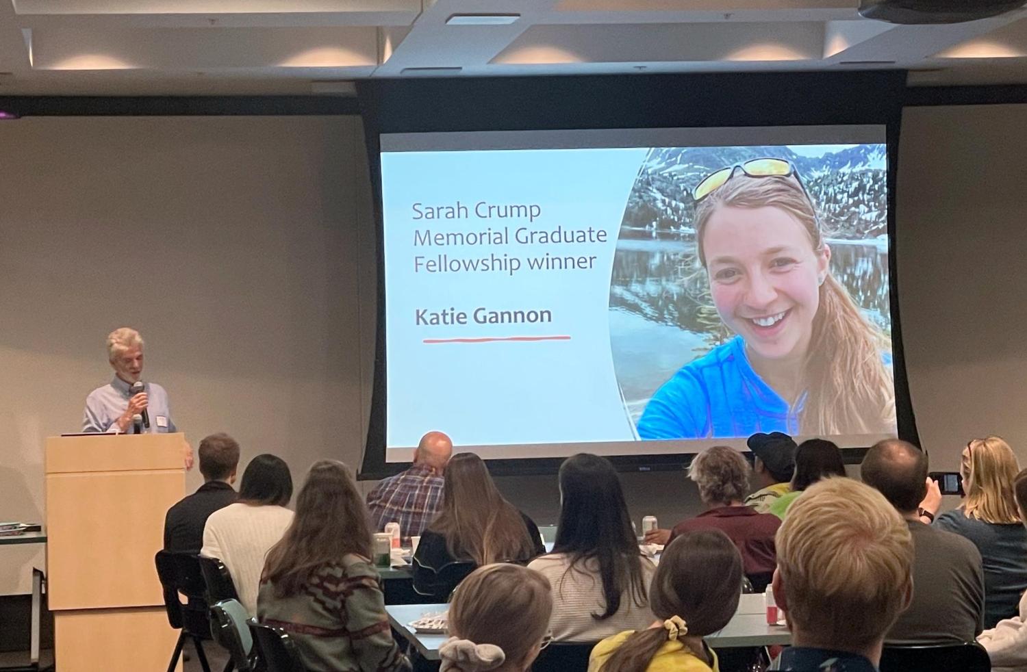 Giff Miller speaking at the 2024 INSTAAR celebration luncheon, with people seated at tables and a projector screen showing Katie Gannon as the winner of the Sarah Crump Graduate Fellowship