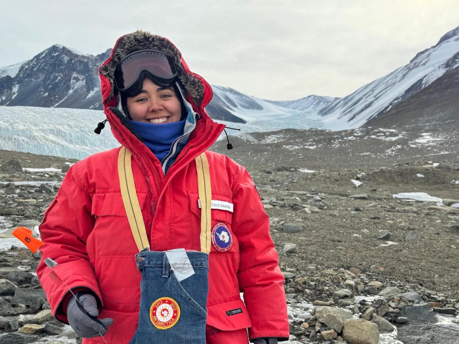 Natalie Aranda, in puffy red parka and ski goggles perched above her eyes, stands on the edge of a rocky Antarctic stream with glaciers and mountains behind her