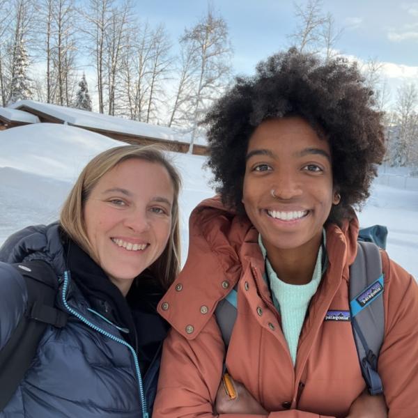 Peyton Thomas and Cassandra Brooks, wearing warm puffy jackets, take a selfie.  Behind them are deep drifts of sparkly snow, covering several buildings.