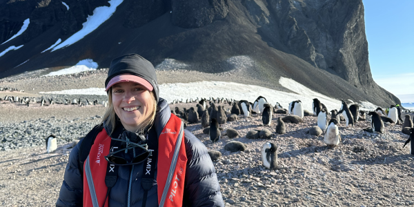 Cassandra Brooks, in warm puffy jacket and brimmed cap, stands near a penguin colony with a dark cliff behind