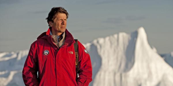 James Balog, in a red jacket with a long-lens camera slung over his shoulder, looks into the distance of an icy landscape.  Behind him is a large jagged iceberg.