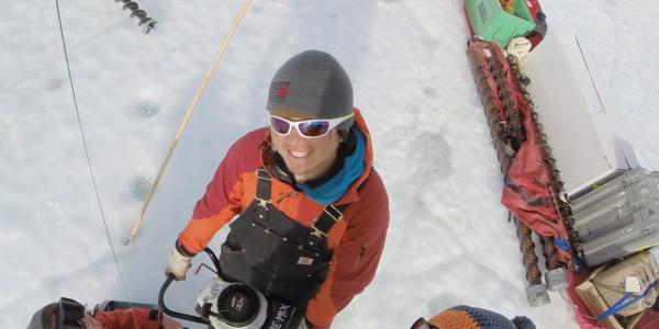 Overhead view of three smiling Antarctic researchers wearing red coats, hats, and sunglasses.  Michael Dyonisius grips a powered ice auger with two hands.  Right next to him are Sarah Shackleton and Bernhard Bereiter.  Photo taken in austral summer of 2015-2016.  Photo overlain with text saying: Meet Michael Dyonisius, INSTAAR postdoc and expert in greenhouse gases, radiocarbon, and ice cores.