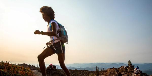 Peyton, a young Black woman wearing a t-shirt, shorts, and backpack, runs along the ridgeline of a mountain trail.