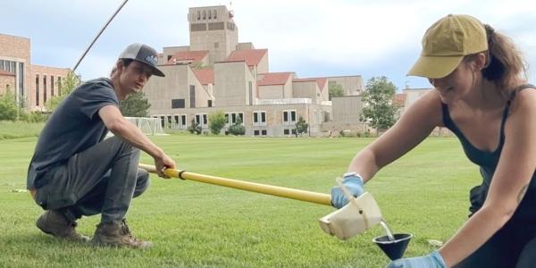 Squatting next to an opened manhole cover on a green campus lawn, a research partner holds a long pole with an attached water sample cup and tips it into a funnelled container held by a rubber-glove wearing Alli Cook. This is one of many water samples that Alli gathered from CU Boulder storm sewers.
