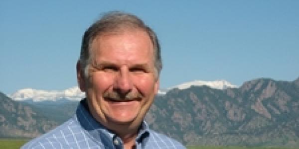 George Aiken, in blue button shirt, stands in sunshine and a grassy field, with the Colorado Front Range in background.