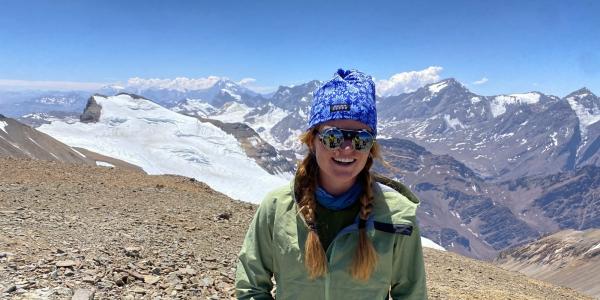 Millie Spencer smiles while standing on the sunny summit of Cerro El Plomo (17,795 feet) in Central Chile.  Behind the rocky summit is a prominent glacier, with a background of mountain peaks with more glaciers and snowfields.  She is wearing a green jacket, blue knit hat, and mirrored glacier glasses