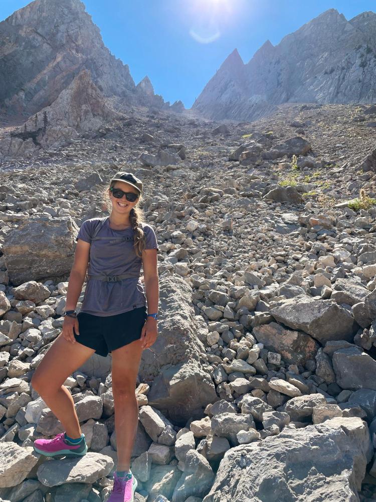Katie Gannon, in running shorts, sunglasses, and baseball cap, poses for a photo on an alpine scree slope with jagged peaks above her.