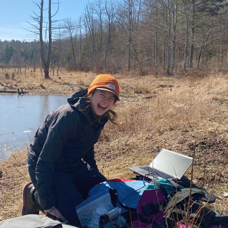 Katie Gannon, in warm black coat and orange knit cap, kneels on a dry grassy lake shoreline while working on a laptop and surrounded by field science gear. Behind her is a mostly deciduous forest without its leaves and a bright blue sky on a cold day.