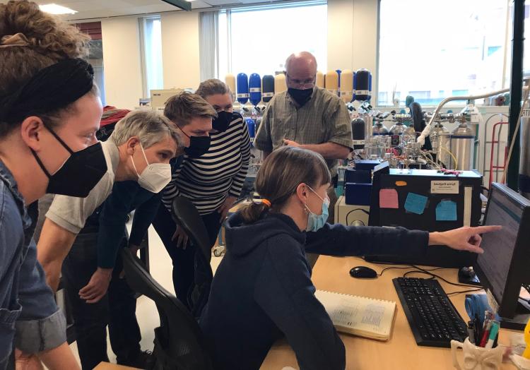 Sitting at a computer, Sylvia demonstrates part of the Stable Isotope measurement process to a group of Finnish visitors closely gathered around her