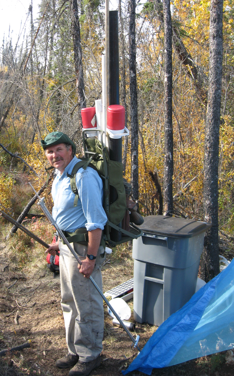 George Aiken gears up for fieldwork, wearing a backpack with long tubes and instruments, and surrounded by other equipment