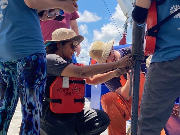 Female researcher with a hat, sunglasses, life jacket and muddy hands kneels down to work a marine sediment core while on the deck of a ship.  She is surrounded by four teammates helping process the core.