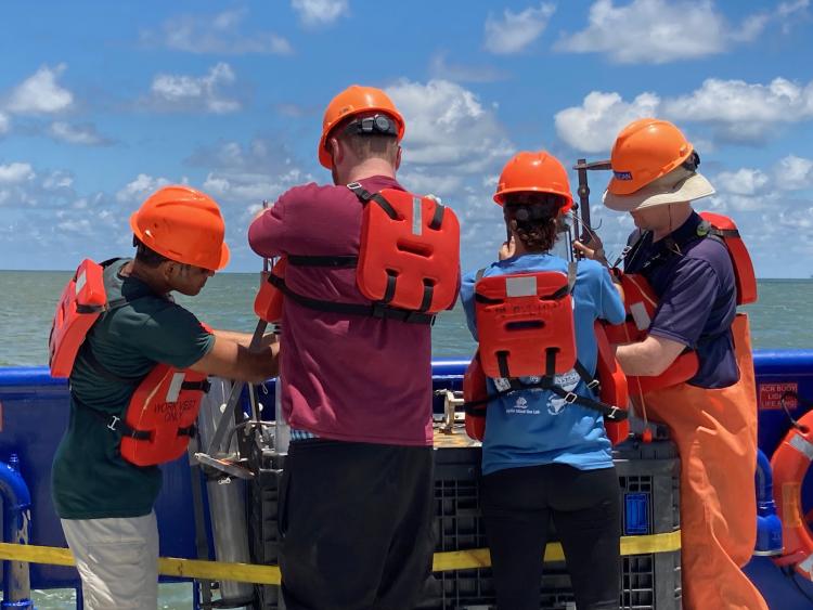 Four researchers in bright orange hard hats and life vests prepare sediment sampling equipment on the deck of a ship in the Gulf of Mexico 