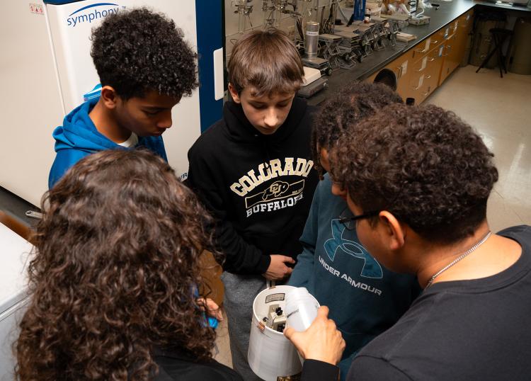 Teens and a scientist huddle around a cylinder of white PVC pipe with metal contraptions inside it. The scientist pours water into the pipe.