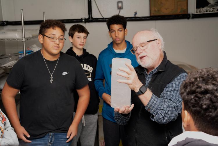 A bald man with white hair and glasses holds up a arm-sized cylinder of pure ice while teens look on with interest