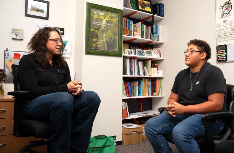 A young hispanic teen in a black shirt talks with a hispanic female scientist, also in a black shirt, sitting across from him