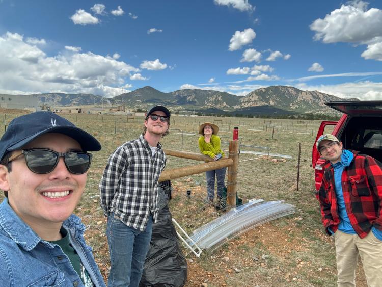 A young man in glasses takes a selfie with three other field scientists in an open pasture with rocky hills in the background at midday