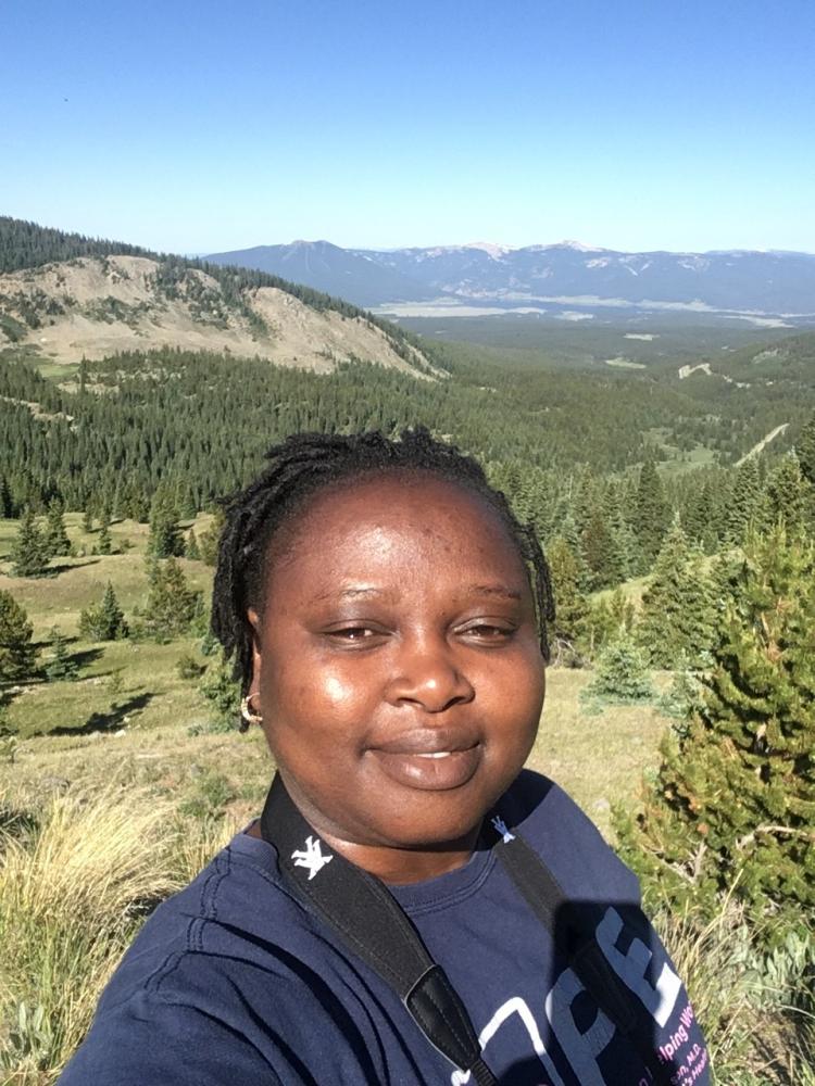 With clear blue skies, Rahila Yilangai takes a summer selfie while doing fieldwork on a forested ridge, with a broad valley below and mountain skyline of Crested Butte Colorado