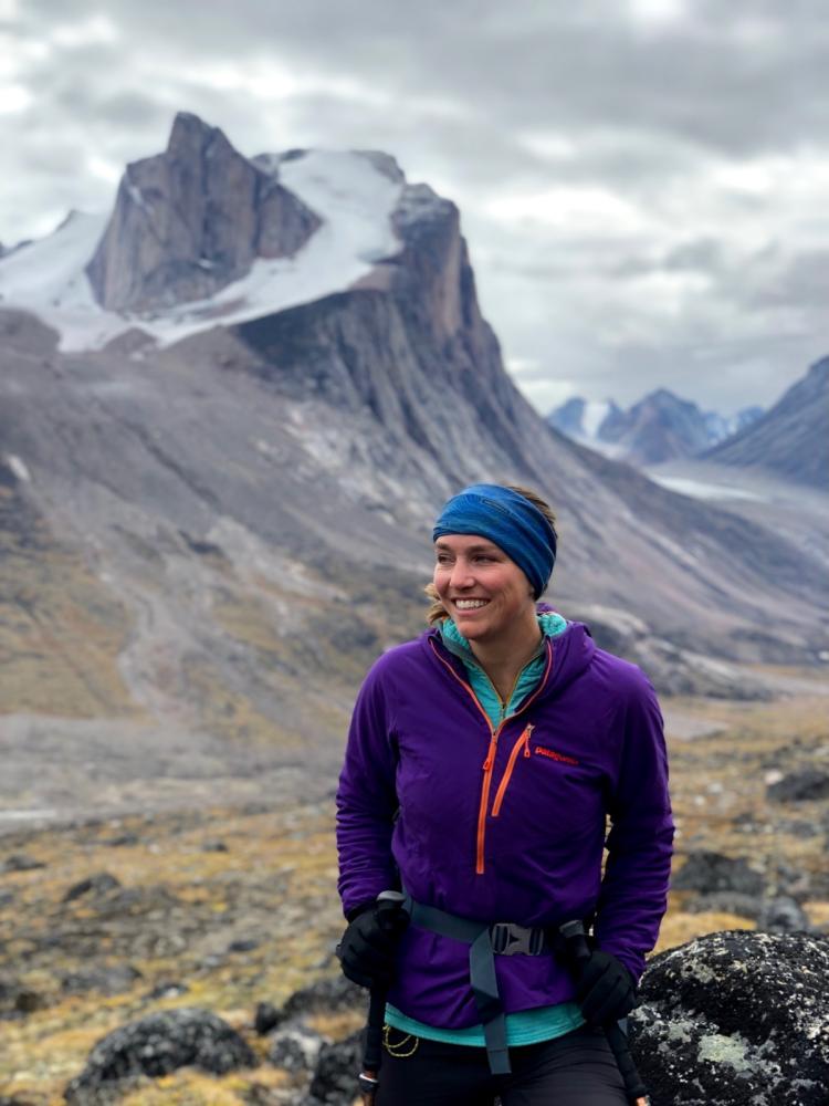 Sarah Crump, in purple coat, stands in front of a dramatic Baffin Island landscape of colorful tundra suddenly broken by towering cliffs.