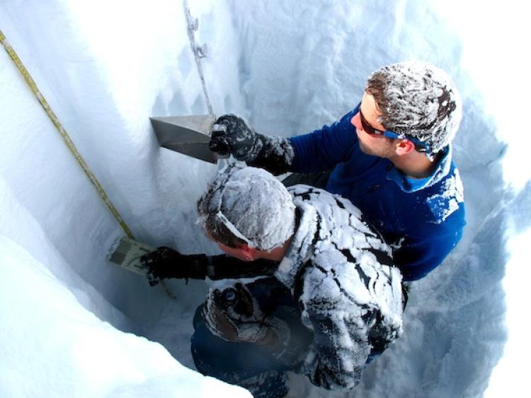 Two hydrology researchers, with snow-covered hair and backs, take snow samples from the sidewall of a snow pit