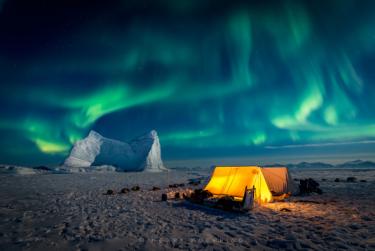 Dramatic aurora looms over several lighted tents camped on sea ice with a frozen-in iceberg behind.  Greenland. Photo by Kerry Koepping