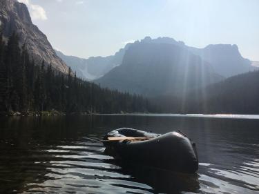 A black inflatable boat sits by itself on a subalpine lake with sun rays and dramatic peaks in the background